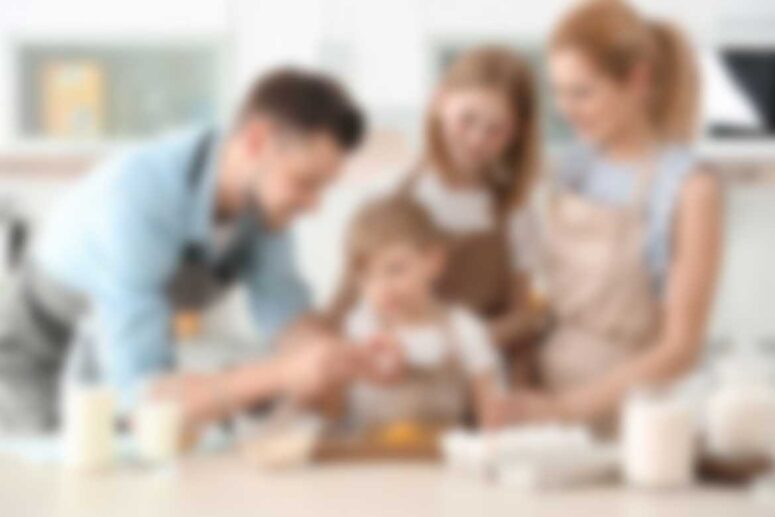 A family of four, including two adults and two children, is gathered in a bright kitchen setting for a baking activity. Donning aprons, they are surrounded by ingredients like eggs and milk on the countertop. Nearby, a Singles Foodies Event flyer lays casually on the table.