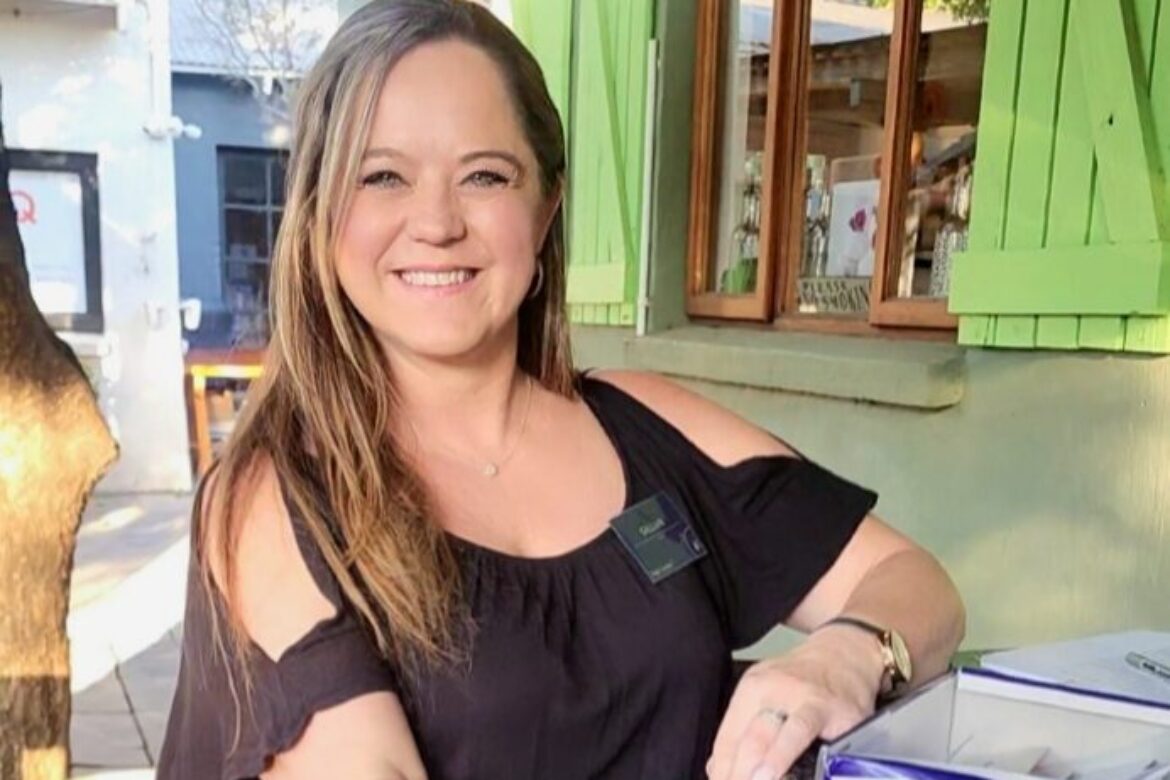 A woman with long hair smiles while wearing a black off-shoulder top. Seated outdoors at a Singles Cooking Club event, she's opening a box labeled "Name Badges." Behind her, green shutters and a window frame the lively setting.
