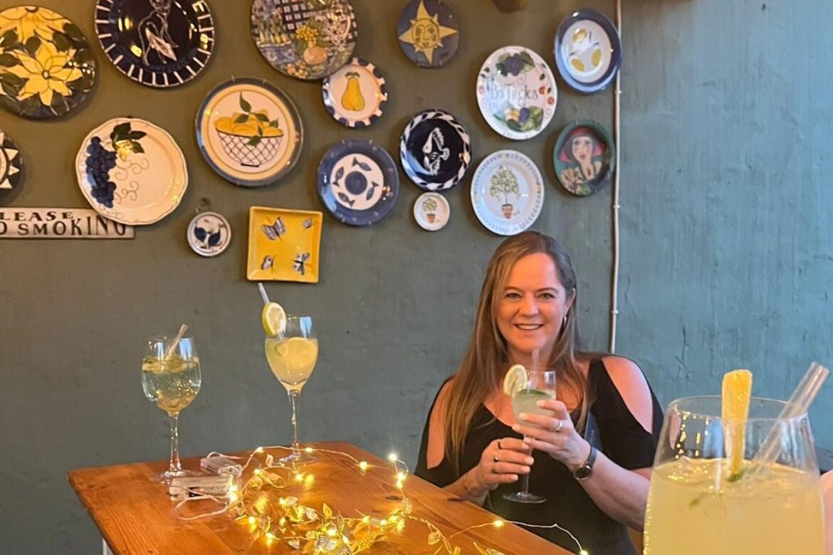 A woman sits at a wooden table with drinks, holding a glass. The wall behind her is adorned with colorful ceramic plates. A string of fairy lights drapes across the table, setting the mood for an evening at the Singles Cooking Club, where culinary talents and connections simmer together.