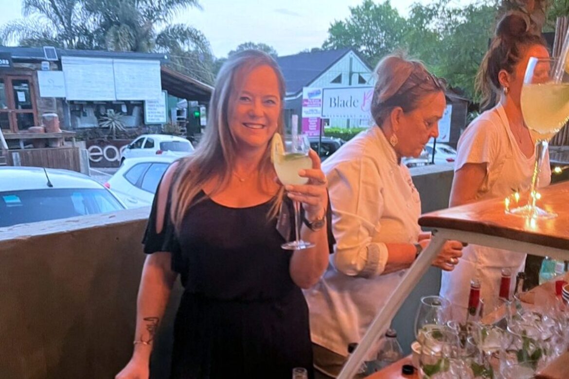 A woman in a black dress is smiling and holding a cocktail glass with a slice of lime. She stands next to a bartender in a white shirt at an outdoor bar, enjoying the vibrant atmosphere of Single Events, with parked cars and trees in the background.