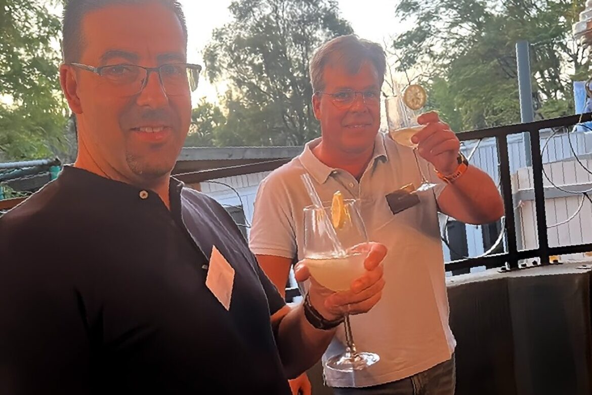Two men from the Singles Cooking Club smile and hold cocktail glasses with lemon slices, standing outdoors. One wears a black shirt and glasses, the other a light polo shirt. Trees and a fence are visible in the background, capturing a relaxed moment at this engaging singles event.