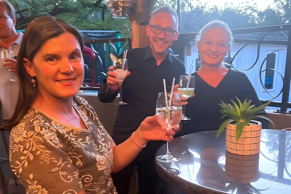Three singles enjoy drinks and smiles at an outdoor gathering. They stand around a table with a small potted plant, as the charm of patio lights and trees enhance this delightful single event.