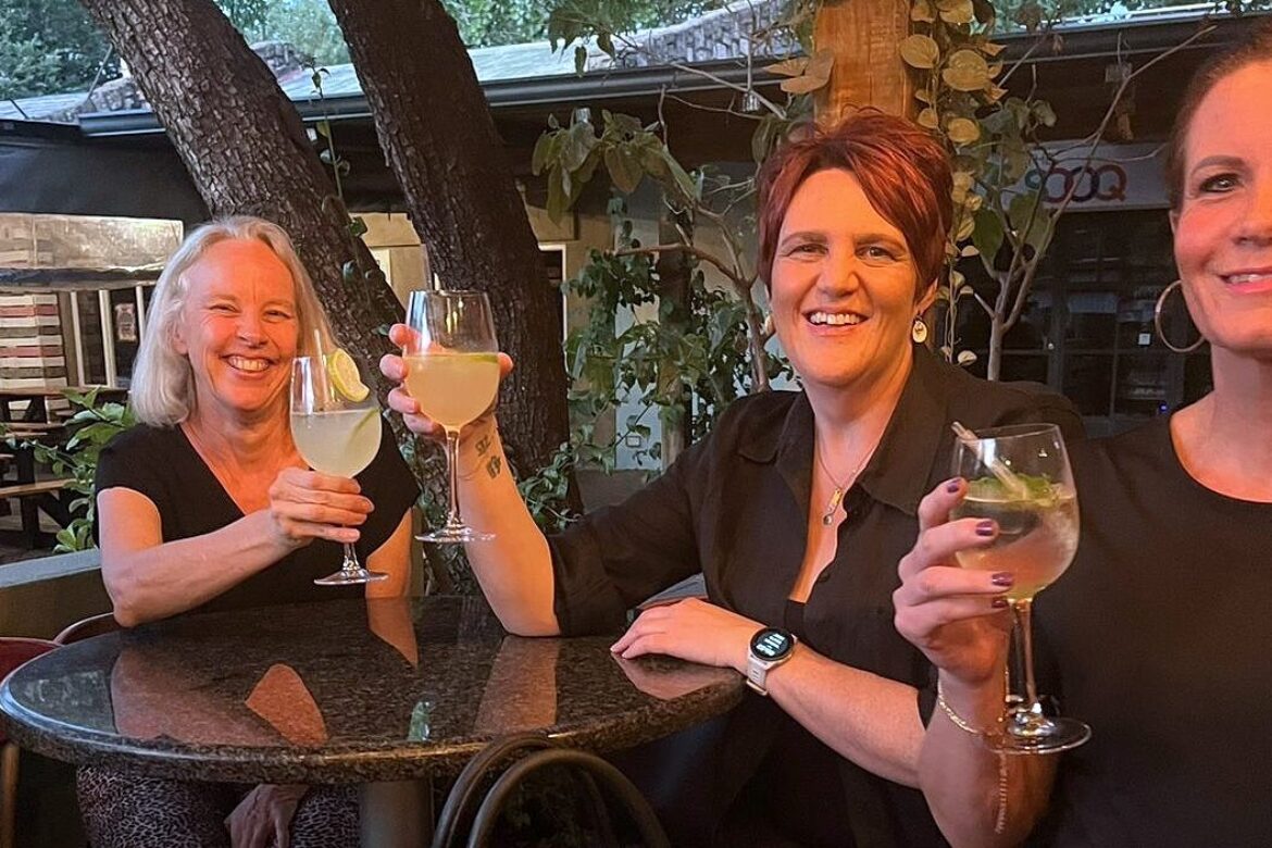 Three women from the Singles Cooking Club sit outdoors at a round table, smiling and holding glasses of light-colored drinks. Trees and greenery in the background create a relaxed, casual atmosphere perfect for socializing at single events.