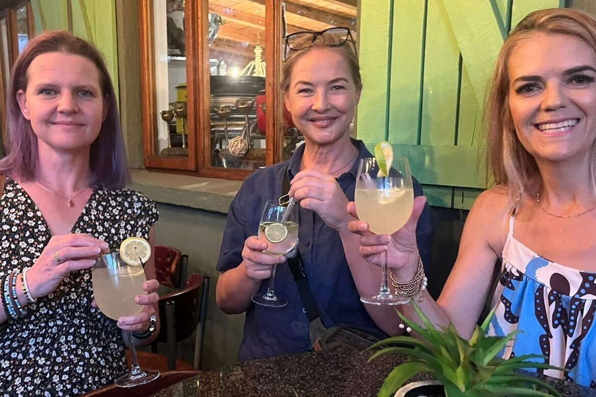 Three women from the Singles Cooking Club smile and hold cocktails with lime and lemon garnishes, standing in a cozy indoor setting with wooden decor and a stone table.