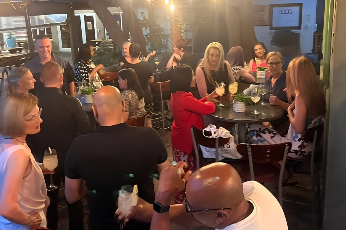 A group of singles is socializing at an outdoor restaurant or bar during the evening. They gather around tables, enjoying drinks and engaging in conversations. Hanging lights and trees create a cozy atmosphere, perfect for mingling and making connections during single events.