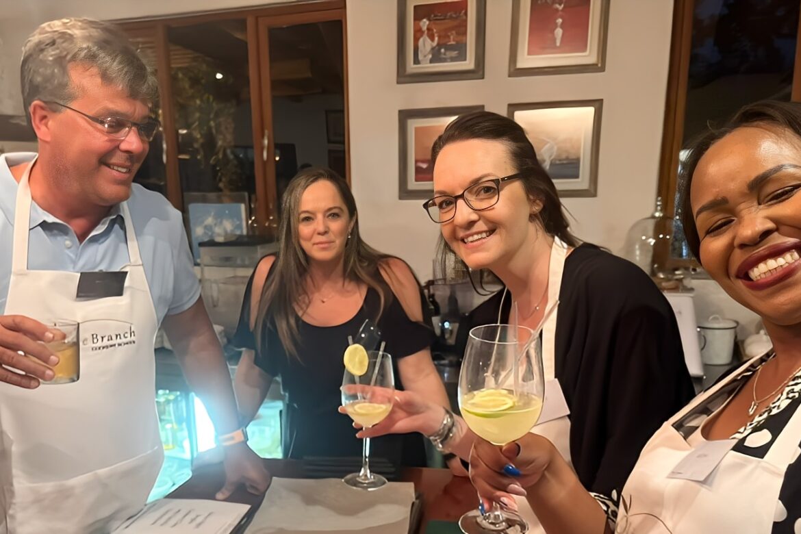 Four people wearing aprons are smiling and holding drinks in a kitchen, part of the lively Singles Cooking Club. Framed pictures hang on the wall in the background, capturing the joy of single events and shared culinary adventures.
