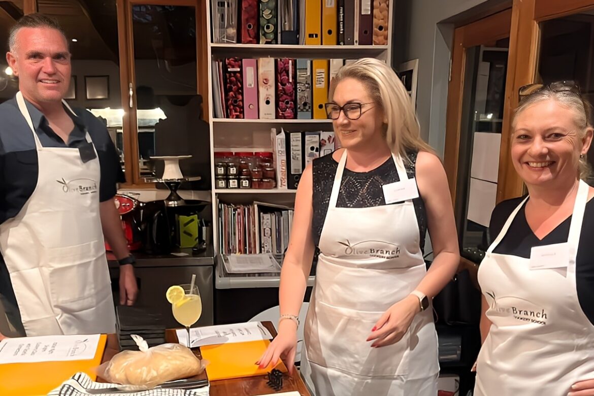 Three people wearing aprons stand in a kitchen, embodying the camaraderie of a Singles Cooking Club. A man and two women smile as they pose around a wooden table with bread and a lemon drink. Shelves filled with binders and jars are in the background, capturing the essence of this lively single event.