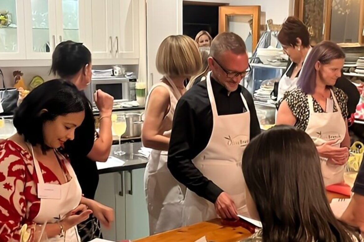 A group of people gather in a kitchen, wearing aprons and engaging joyfully in a Singles Cooking Club event. They appear to be preparing food together, with various kitchen utensils and ingredients scattered across the counter around them.