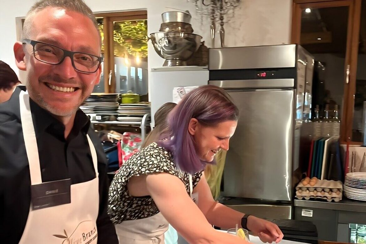 A man wearing glasses and a white apron smiles at the camera. Next to him, a woman with purple hair, wearing a black and white patterned top, focuses on preparing food. They are in a well-equipped kitchen at the Singles Cooking Club, surrounded by stacked dishes and a refrigerator.