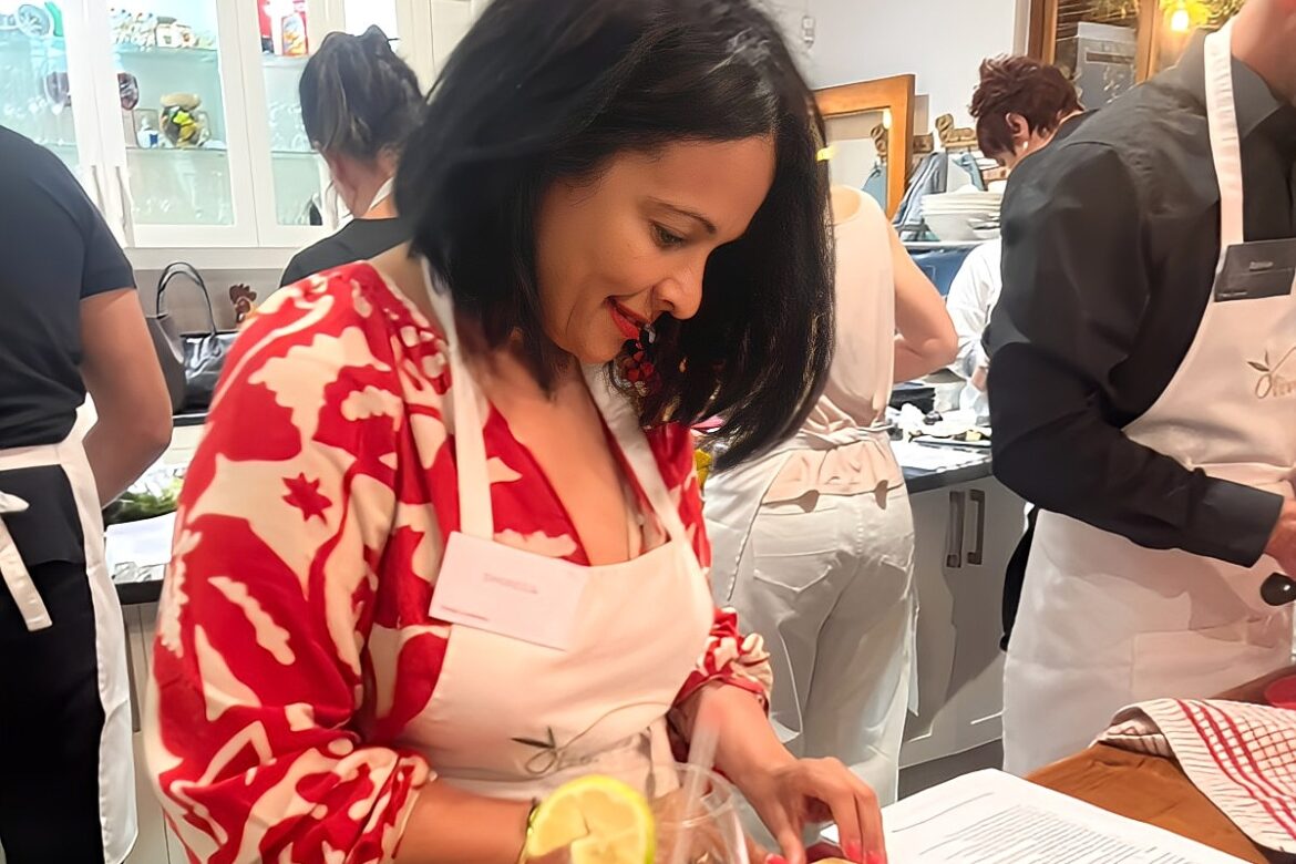 In the bustling Singles Cooking Club kitchen, a woman in a red and white patterned shirt chops vegetables on a cutting board, her apron dusted with flour. A glass with a lemon garnish sits elegantly in the foreground, while other singles join in the culinary fun around her.