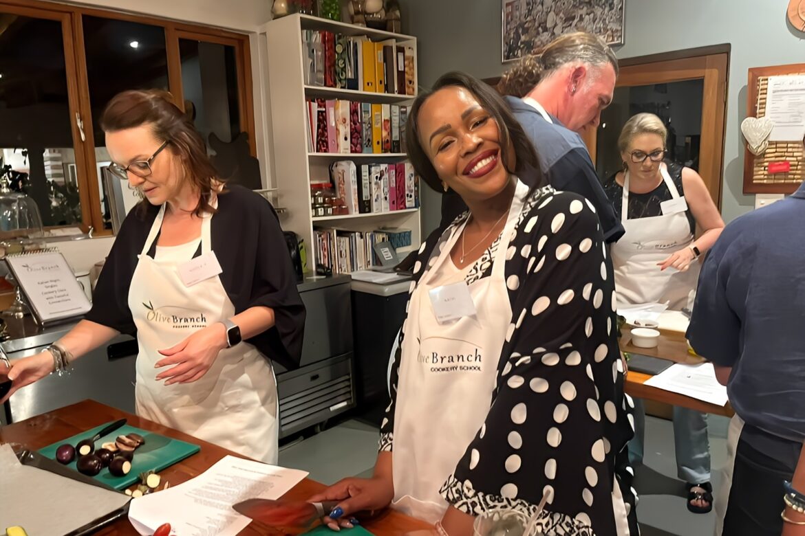 People are gathered around a kitchen island wearing aprons at a Singles Cooking Club event. The focus is on a smiling person in the foreground next to chopped vegetables. Books and kitchen items are visible in the background, adding to the lively and inviting atmosphere.