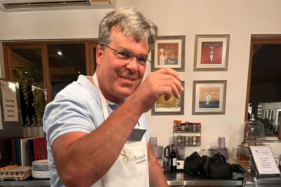 A smiling man in a white apron raises a glass in a bright indoor setting, embodying the spirit of the Singles Cooking Club. Behind him, shelves display framed pictures, spice jars, and a glass cake stand, creating an inviting atmosphere for those single events that promise fun and connection.