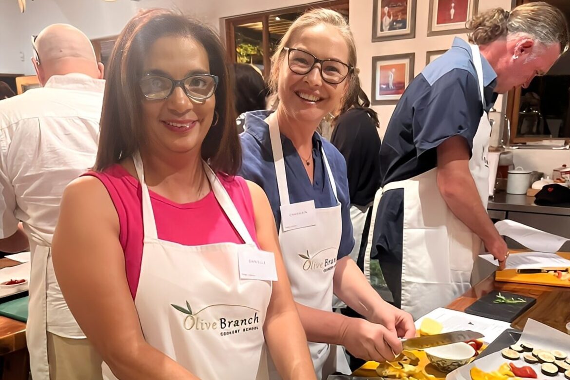 Two smiling women in aprons stand at a kitchen counter, preparing food for the Singles Cooking Club. They are surrounded by ingredients and cooking utensils, while others engaged in cooking activities fill the lively and friendly atmosphere.