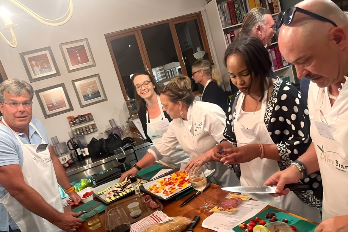 A lively Singles Cooking Club gathers around the kitchen table, aprons on, preparing food. Colorful ingredients like diced fruits and vegetables are spread out as everyone collaborates cheerfully, creating both delicious dishes and new connections.