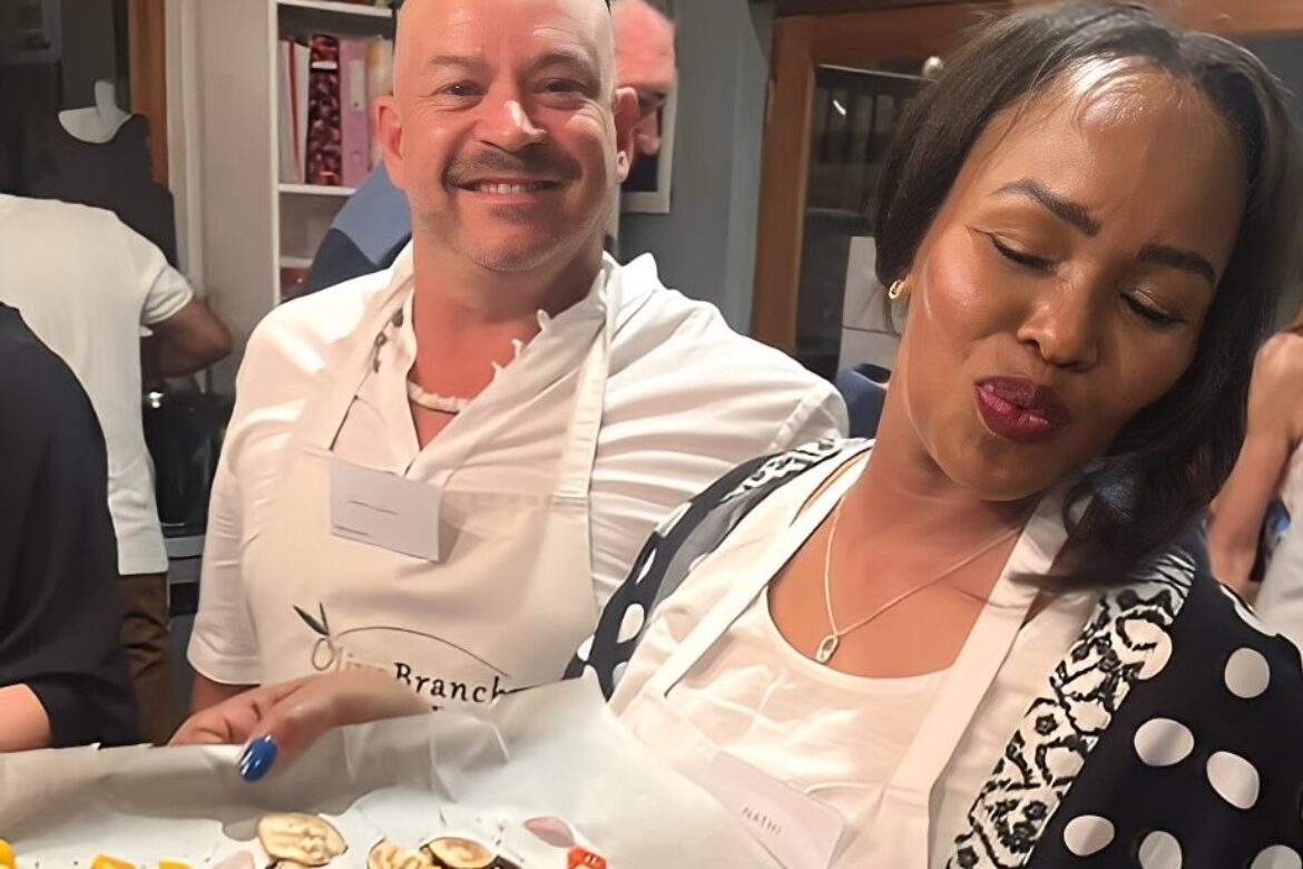 Two people wearing aprons stand in a kitchen at the Singles Cooking Club event. The man smiles at the camera, while the woman looks down, carefully holding a tray of colorful diced vegetables and crackers. Others are mingling joyfully in the background.