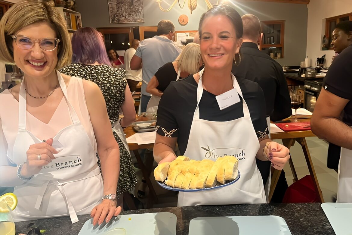Two women wearing aprons stand in a lively kitchen, part of the Singles Cooking Club. One holds a plate of sliced bread, smiling warmly. In the background, singles are joyfully engaged in various cooking activities, creating a vibrant and inclusive atmosphere.