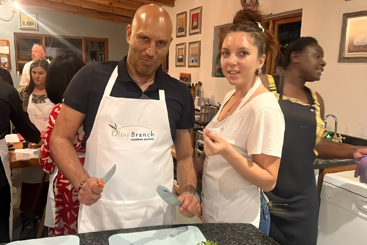 A man and woman in aprons participate in the Singles Cooking Club at Olive Branch. The man holds a pizza cutter and poses playfully, while the woman looks at the camera. Other participants are in the background. The setting is cozy and well-lit, perfect for single events.