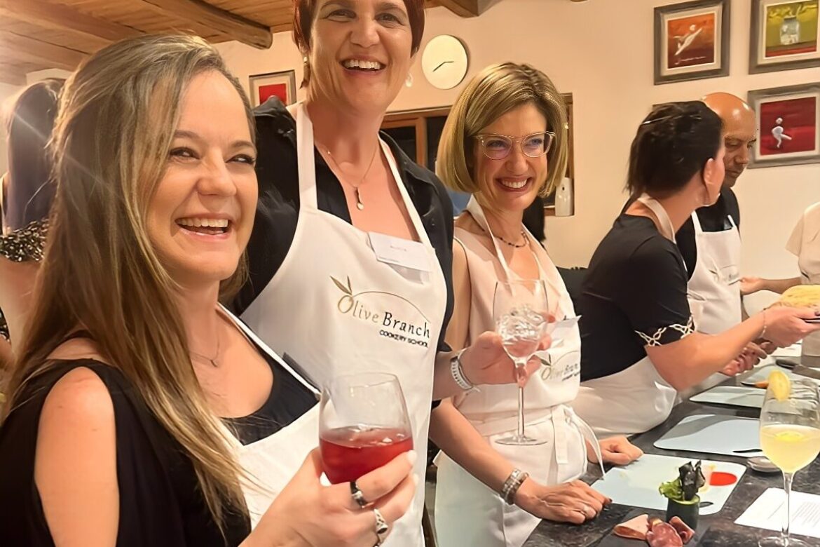 A group of people in a kitchen, part of the Singles Cooking Club, wear aprons and smile while holding glasses of wine and cocktails. They stand behind a counter with plates of food, participating in a lively cooking class. The room features a wooden ceiling and framed pictures on the wall.