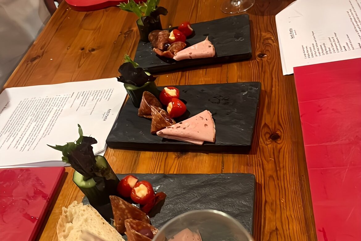 A wooden table set for a Singles Cooking Club class features slate boards with cured meats, cheeses, bread, and stuffed peppers. Red cutting boards, kitchen utensils, and recipes are scattered nearby. A cocktail with a cucumber garnish is in the foreground.