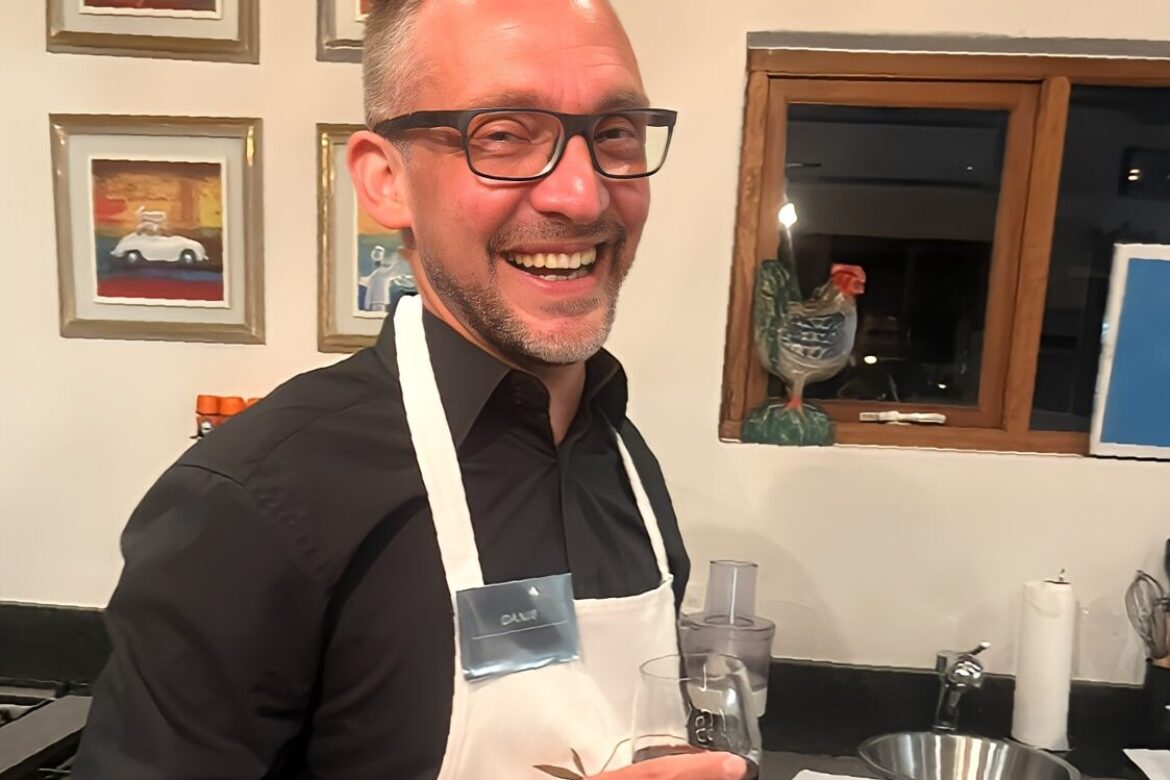 A man wearing glasses and an apron stands in a kitchen, part of the Singles Cooking Club, holding a drink and smiling at the camera. Artwork frames and a rooster sculpture grace the background, along with a kitchen counter and window, setting the stage for this delightful single event.