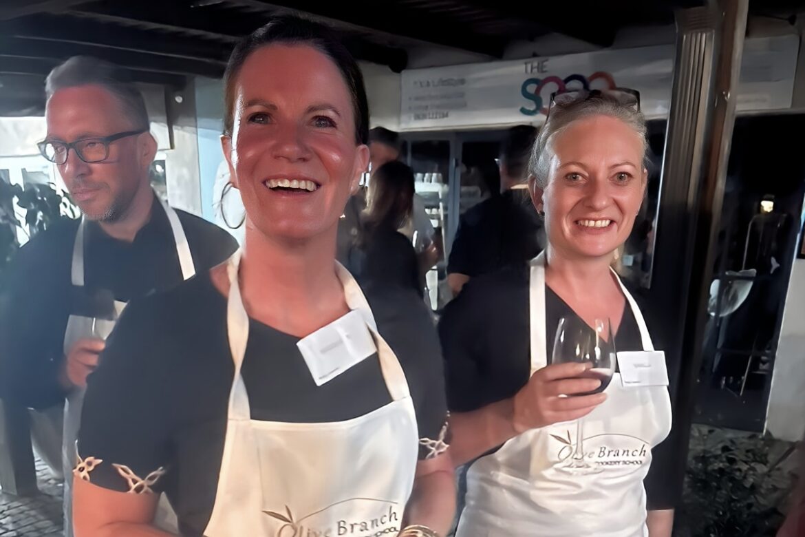 Three people wearing aprons with name tags are smiling at a Singles Cooking Club event. One holds a glass of red wine, adding to the casual, social atmosphere indoors.