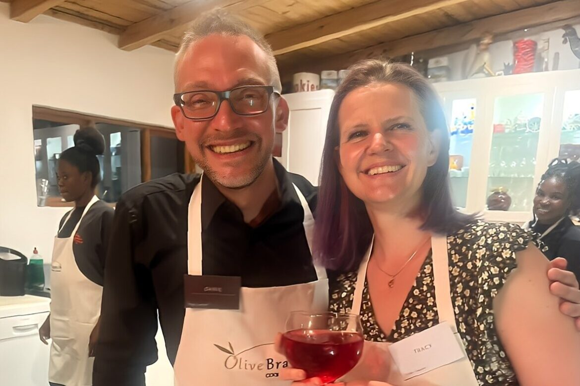 A man and a woman are smiling and standing close together in a kitchen, part of the Singles Cooking Club. Both wear aprons with name tags, and the woman is holding a glass of red liquid. Other singles are visible in the background, also in aprons, enjoying the lively cooking event.