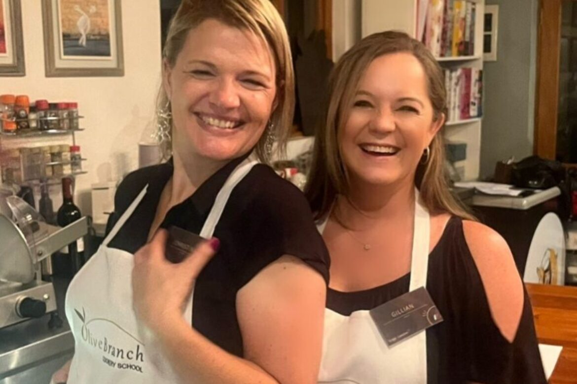 Two women smiling and wearing white aprons stand together in a cozy kitchen setting, part of the Singles Cooking Club. Shelves filled with cookbooks and spices are visible in the background, creating a warm atmosphere perfect for single events.