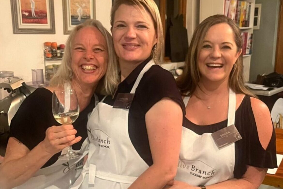 Three women wearing aprons are smiling and standing together in a kitchen, part of the lively Singles Cooking Club. The woman on the left is holding a glass of white wine. Behind them are framed pictures and shelves filled with books and containers.