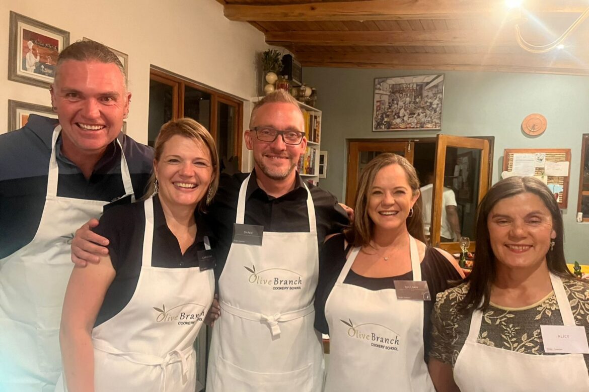 A group of five singles wearing aprons smile at the camera. They are indoors, standing close together, and appear to be participating in a Singles Cooking Club class. There are pictures on the walls and a wooden ceiling in the background.