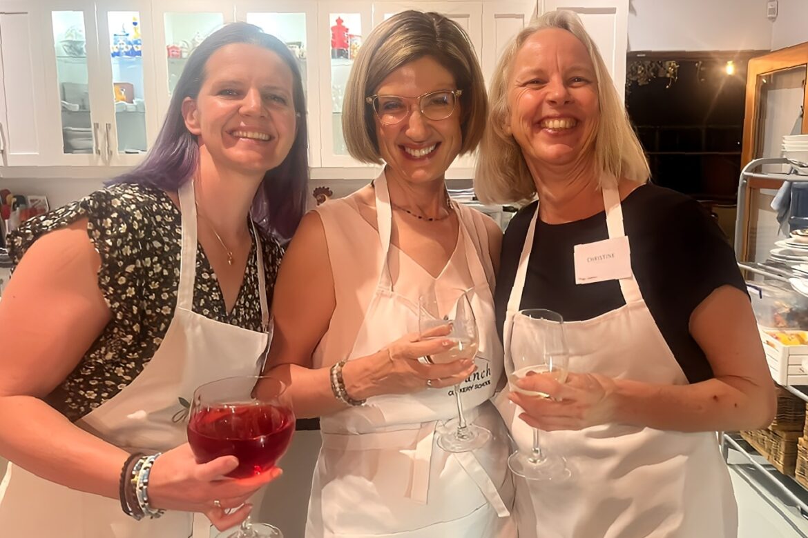 Three women wearing aprons stand in a kitchen, smiling and holding glasses. The kitchen, with its white cabinets and various utensils, is the perfect backdrop for this Singles Cooking Club event. They appear to be enjoying a delightful social gathering or cooking session.