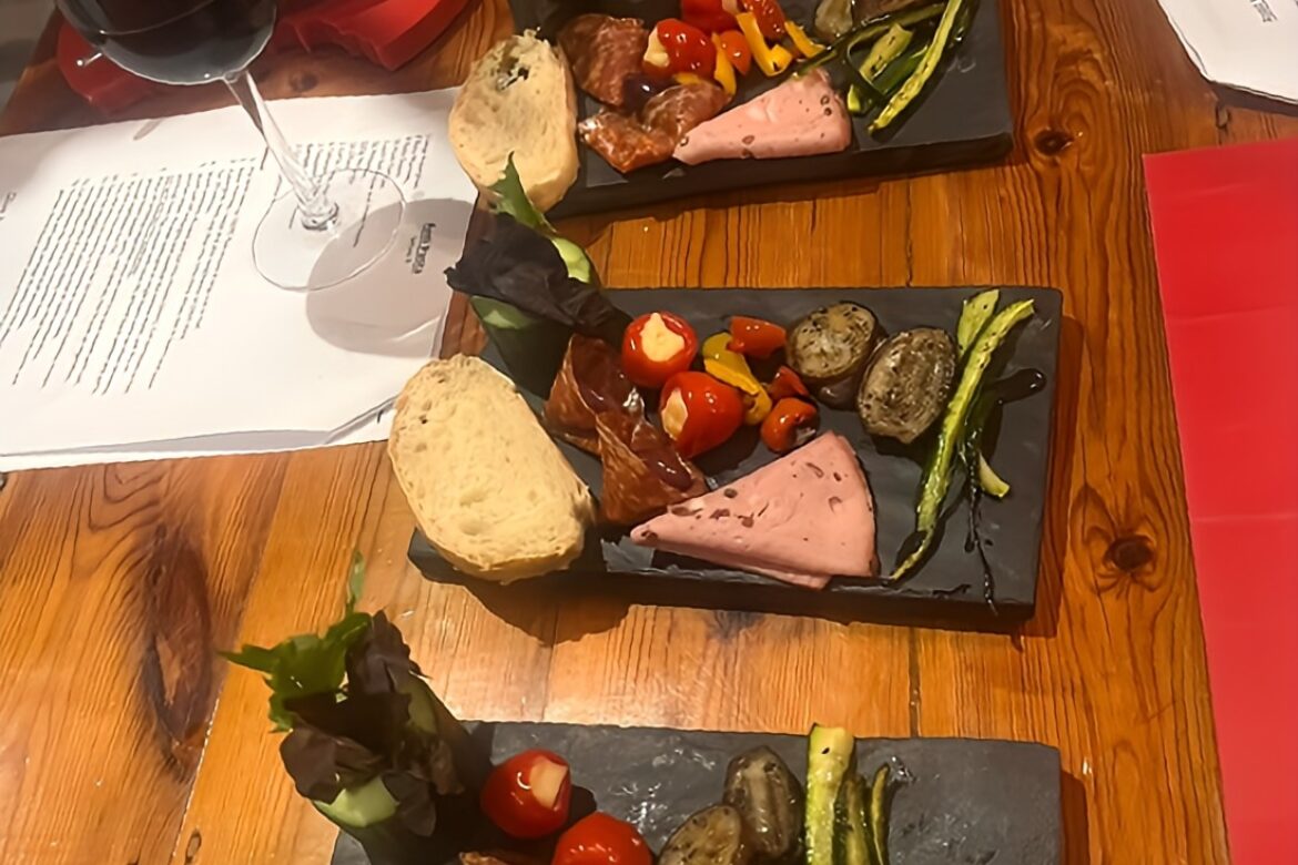 At a Singles Cooking Club soirée, three slate platters display a tempting array of charcuterie—cured meats, cheese, bread slices, cherry tomatoes, asparagus, and pickles. A glass of red wine rests on the wooden table alongside scattered papers and a partially visible plate in the background.