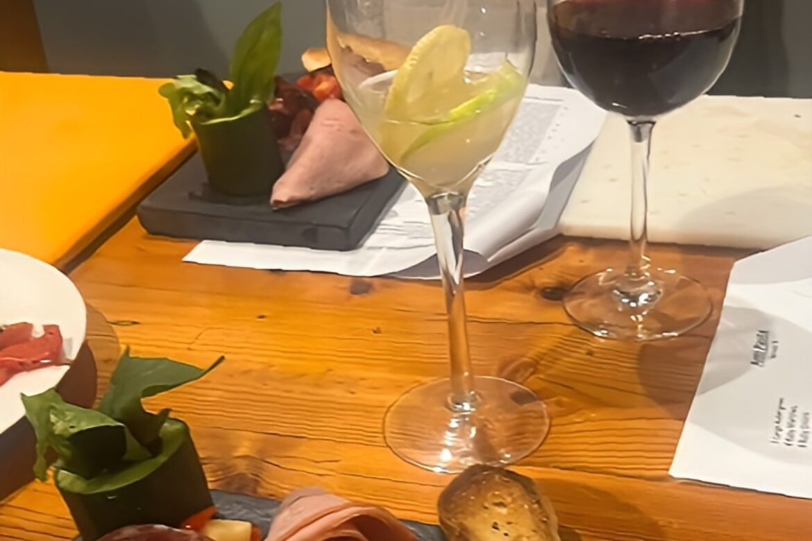 A wooden table set for the Singles Cooking Club boasts a plate of assorted cold cuts and vegetables, garnished with breadsticks. Two wine glasses sit nearby—one with white wine and lemon slices, the other with red wine. Papers are scattered in the background, hinting at an evening of single events planning.