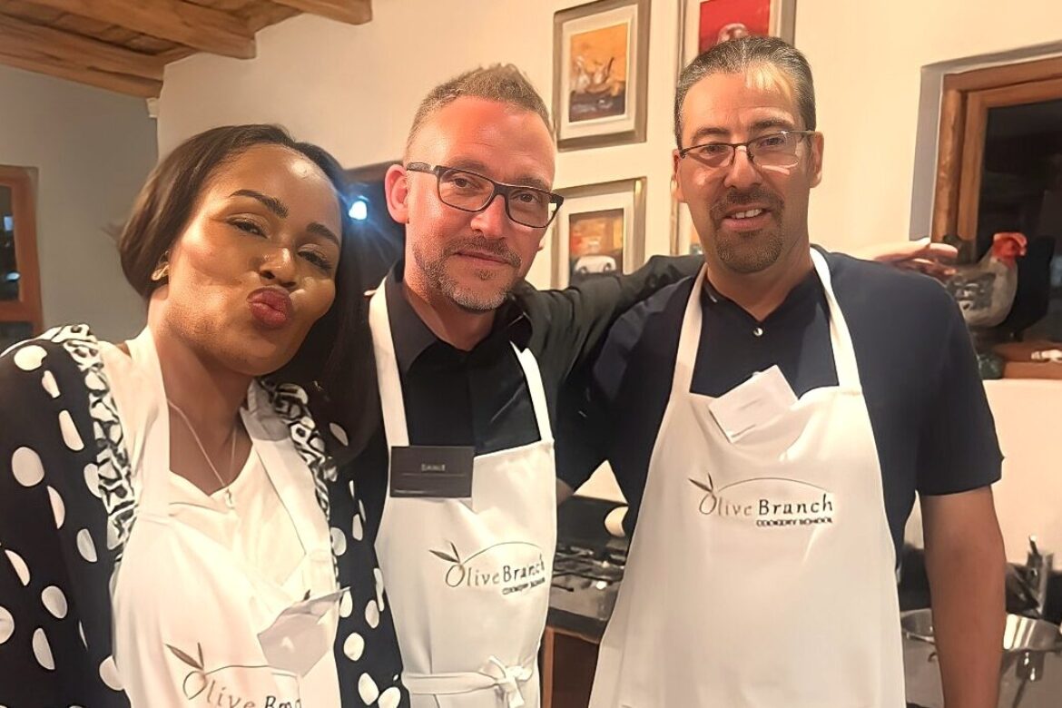 Three people in aprons at the Singles Cooking Club are posing for a photo. The person on the left is smiling with puckered lips, the person in the middle is wearing glasses, and the person on the right is also smiling. Kitchen decor adds charm to this lively single event backdrop.