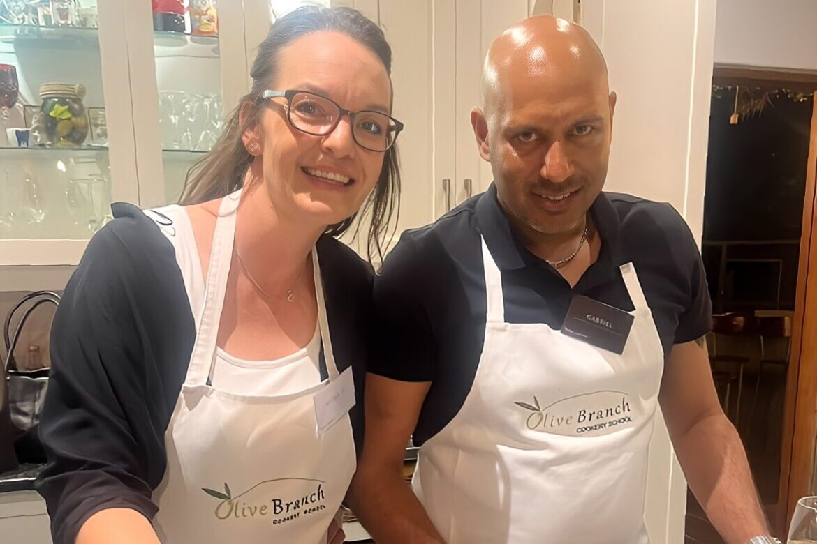 Two people wearing aprons that read Olive Branch Cooking School are smiling in a kitchen, enjoying a Singles Cooking Club event. They appear to be preparing food together, with cabinets and kitchenware visible in the background.