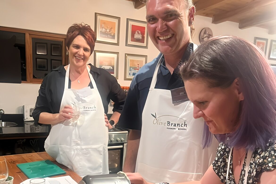 Three people are in a kitchen wearing aprons, part of the Singles Cooking Club. Two are smiling at the camera, while the third is using a mixing machine. The room features a wooden ceiling and framed pictures on the walls, capturing the lively atmosphere of a cooking class for singles.