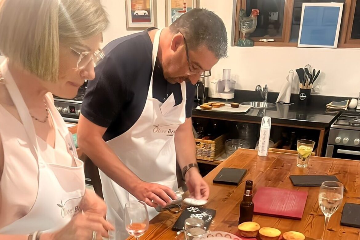Two people in aprons from the Singles Cooking Club are decorating small cakes at a wooden kitchen table. Various kitchen tools, a few drinks, and baked goods are on the table. They are focused on their work, indicating a casual and enjoyable baking session for this single event.