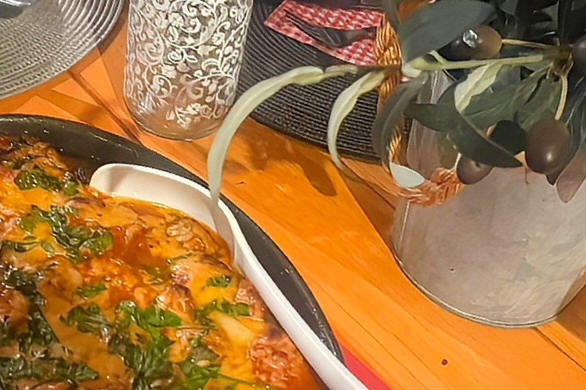 A baked dish topped with melted cheese and herbs sits on a wooden table, perfect for a Singles Cooking Club gathering. Nearby are a decorative bottle, a plant in a metal pot, some red and white napkins, and a white serving spoon.
