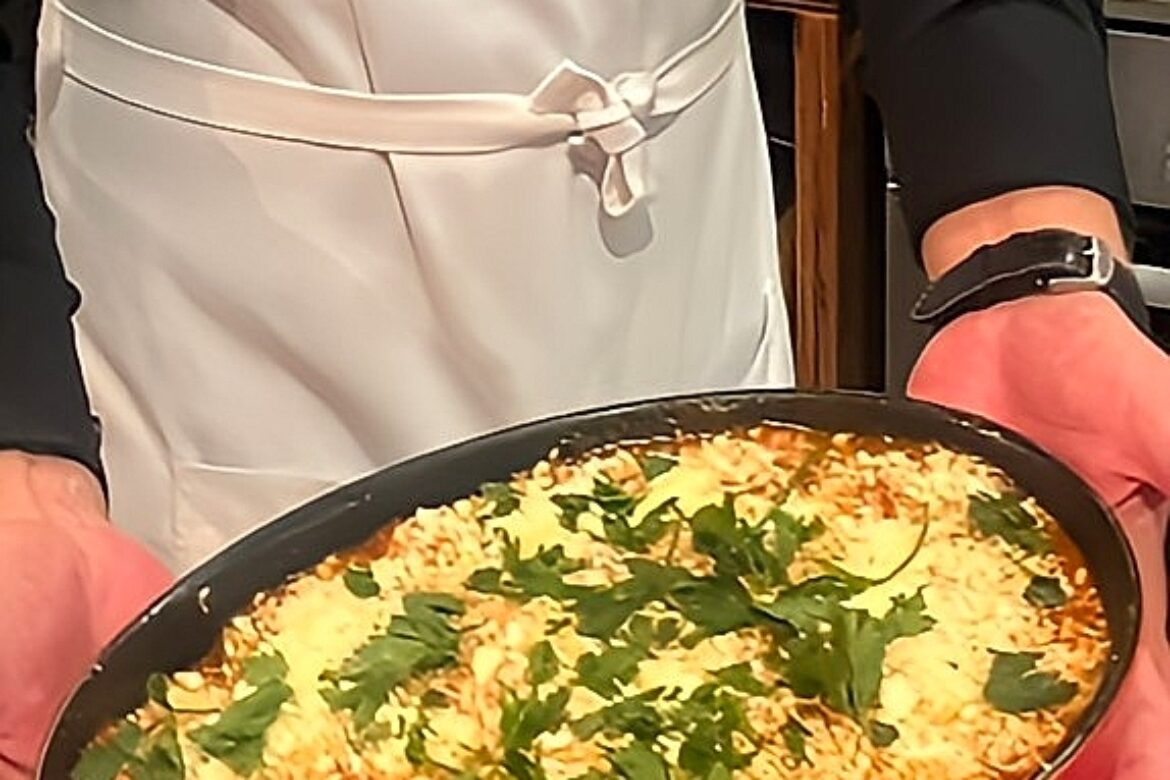 A person wearing a white apron from the Singles Cooking Club holds a red baking dish filled with a creamy, cooked dish topped with melted cheese and garnished with green herbs. The apron proudly displays Olive Branch Cookery School.
