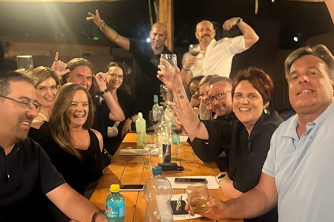 A group of people from the Singles Cooking Club sit around a wooden table, smiling and raising their glasses in a celebratory toast. They enjoy an outdoor social gathering under the night sky, surrounded by various bottles and glasses.