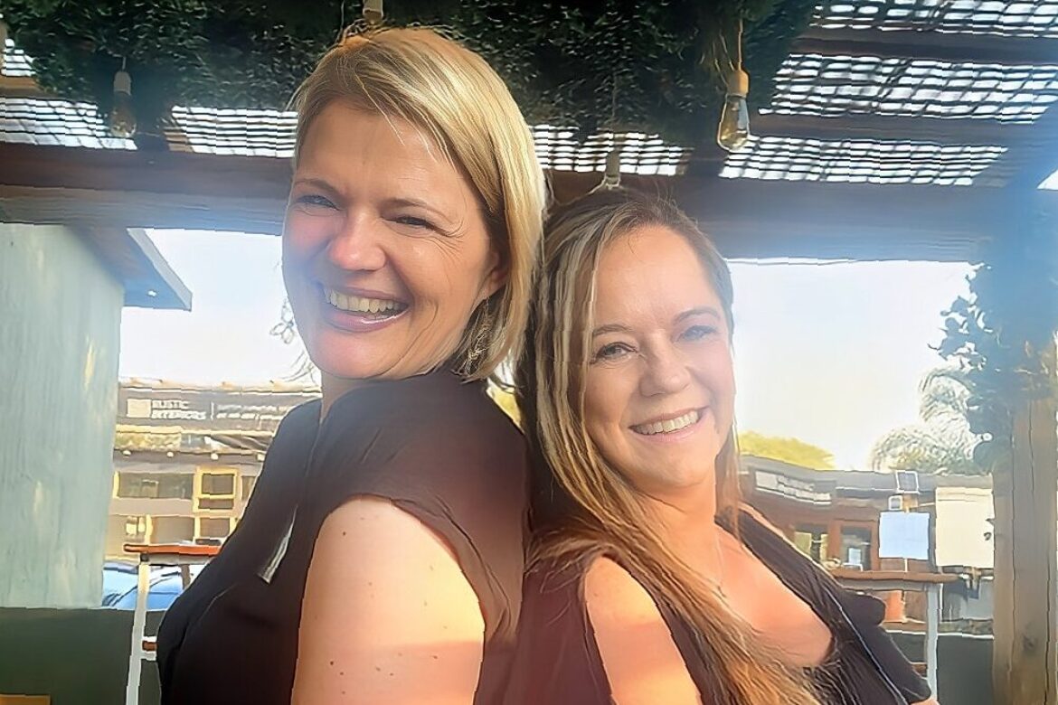 Two women, radiating joy, stand back-to-back under a pergola adorned with greenery and twinkling lights, smiling for the camera. They're dressed in black tops and basking in the sunny day at a Singles Cooking Club event.