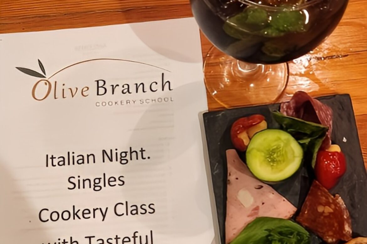 A wooden table features a flyer for Olive Branch Cookery School's Italian Night Singles Cookery Class, perfect for charming singles events. Beside it, a glass of iced beverage and a slate platter with assorted deli meats, cheese, bread, and salad create an inviting setting.