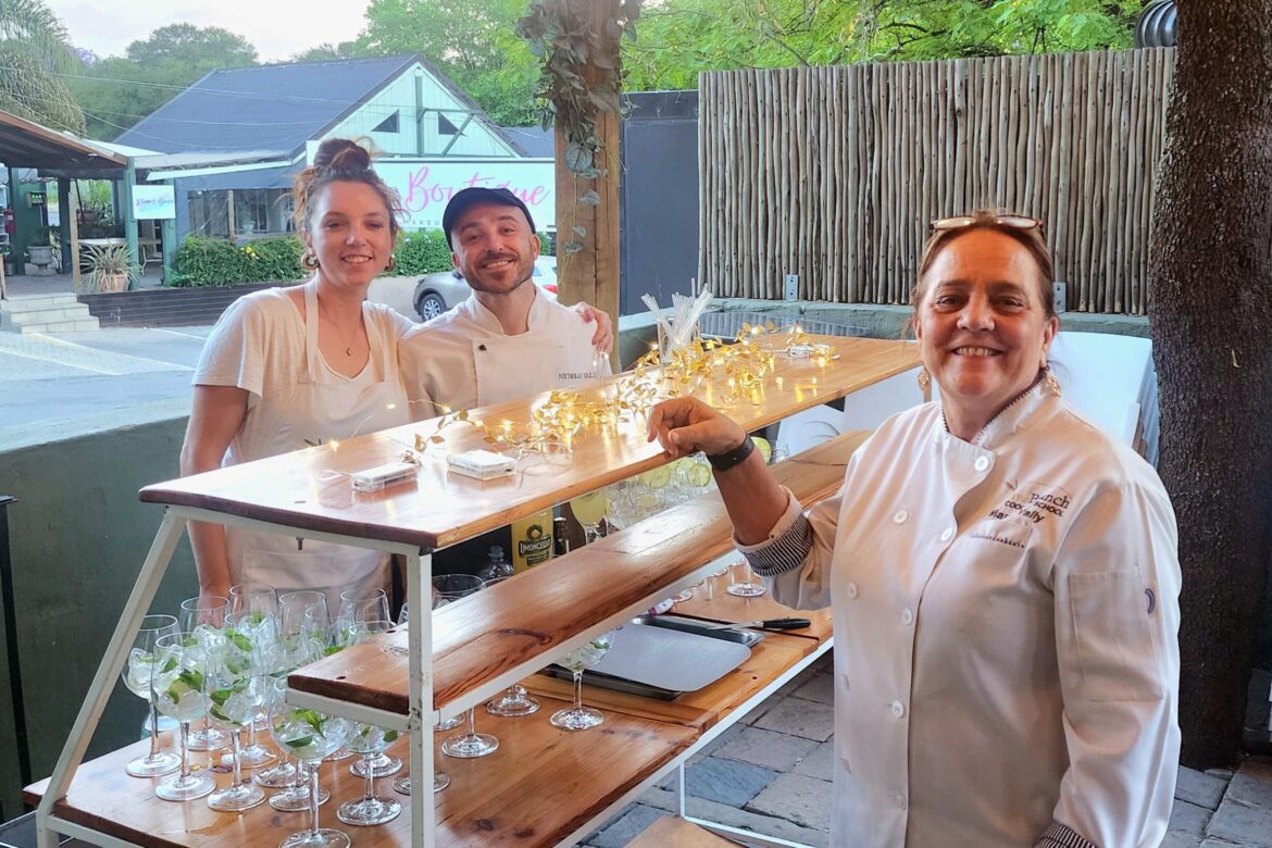 Three chefs from the Singles Cooking Club stand behind a wooden outdoor bar, surrounded by empty glasses and twinkling lights. They smile warmly in a relaxed, lush setting with greenery and buildings in the background, showcasing the joy of connecting through culinary adventures.