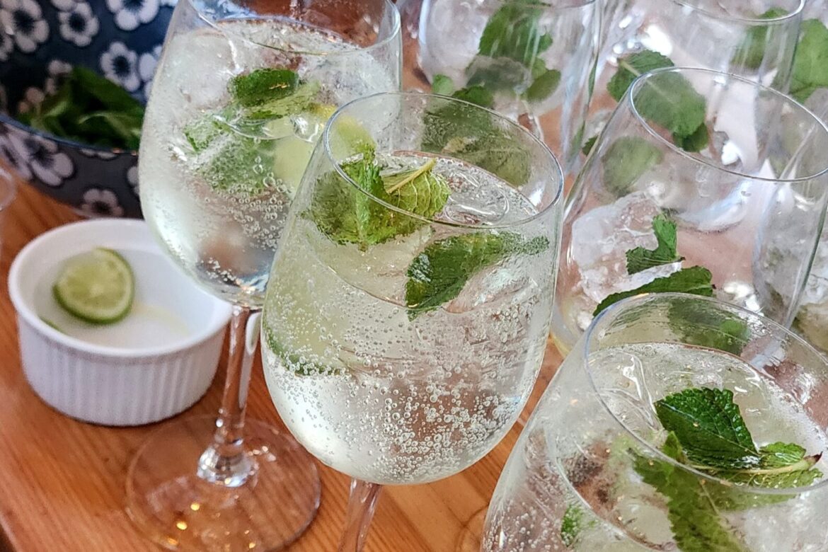 Several glasses of sparkling water with mint leaves and lime slices are arranged on a wooden counter, perfect for a Singles Cooking Club gathering. A small white dish with lime slices sits nearby, and a flower-patterned bowl adds charm to the background ambiance.