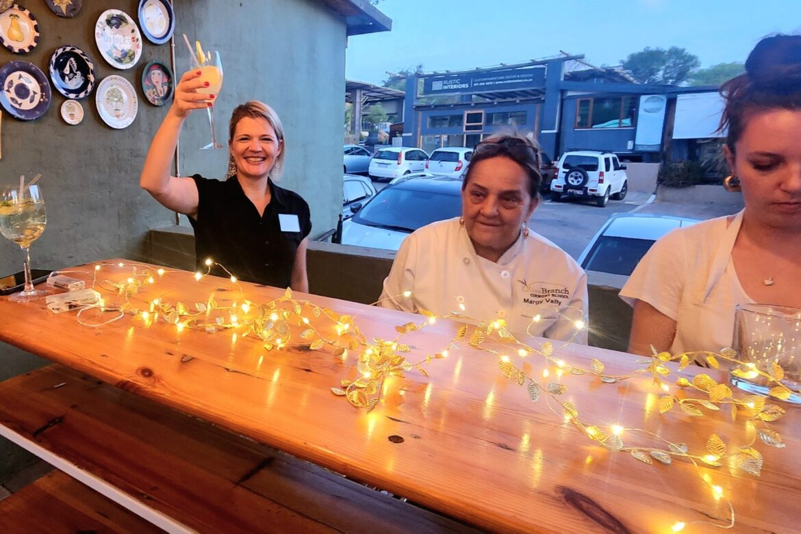 Three women are gathered around a wooden bar adorned with string lights. One woman on the left is smiling and raising a glass, while the woman in the middle wears a chef's jacket, hinting at a lively Singles Cooking Club event. Plates decorate the wall behind them, with cars visible outside.