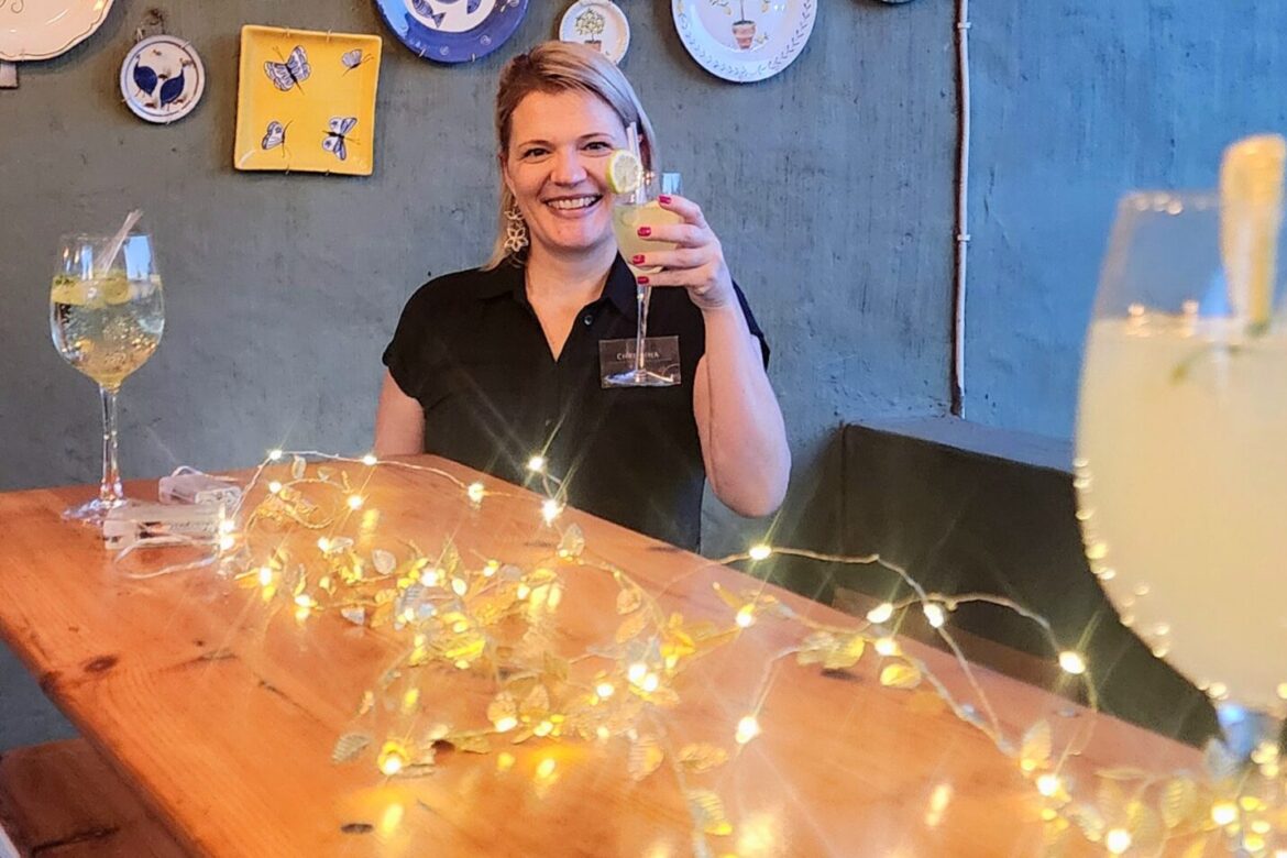 A person in a black shirt holds a cocktail glass while sitting at a wooden table adorned with string lights, enjoying the cozy and cheerful ambiance of the Singles Cooking Club. Decorative plates hang on the wall behind them, setting the perfect scene for social single events.