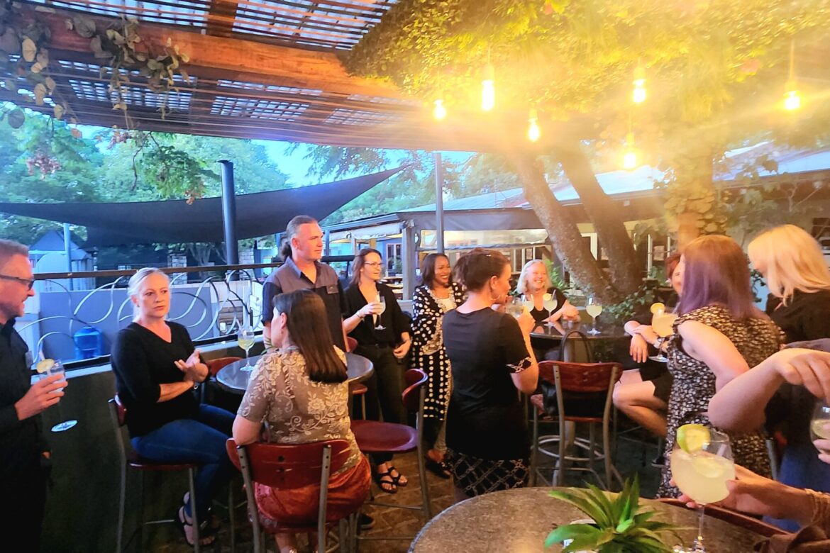 A group of singles is socializing at an outdoor bar with string lights overhead. They gather around high tables, enjoying drinks and vibrant conversations. The setting, perfect for single events, has a rustic vibe with lush greenery and a wooden pergola completing the charming scene.