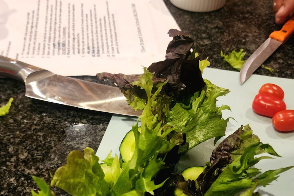 A kitchen counter displays an array of sliced cucumbers and leafy greens meticulously arranged in stacks. Cherry tomatoes sit near a knife, ready for action. Papers with text are scattered in the background, while hands from the Singles Cooking Club actively prepare a vibrant meal.