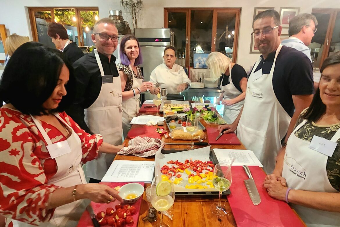A group of singles in aprons participate in a lively cooking class, surrounding a table brimming with fresh ingredients like vegetables and fruits. As part of the Singles Cooking Club, they enjoy preparing a dish with guidance from the chef, creating a friendly and casual atmosphere.