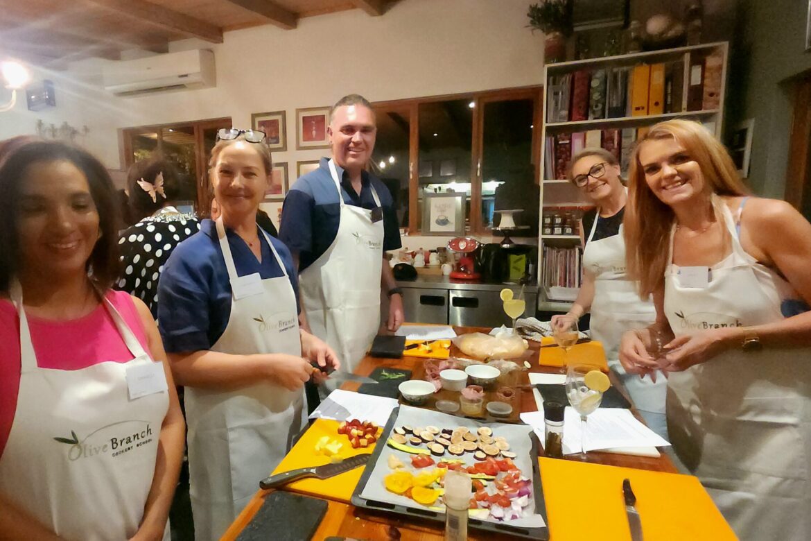 A group of five singles, wearing aprons, are gathered around a kitchen island with ingredients and recipes. They're smiling as they enjoy a Singles Cooking Club class. In the background, shelves with jars and cooking utensils complete the cozy scene.