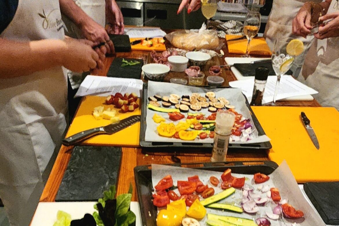 People wearing aprons are gathered around a kitchen counter, preparing food at the Singles Cooking Club. Various vegetables and ingredients are neatly arranged on trays, with beverages in the foreground. The scene suggests a lively cooking class or group cooking activity.